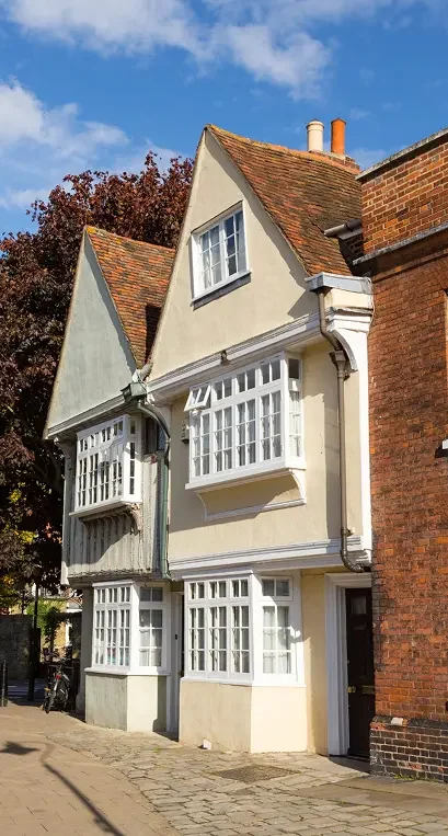 A historic English townhouse with white-framed bay windows, pale stucco walls, and a steeply pitched tiled roof stands beside a red brick building—an inviting scene for those considering First-Time Buyer Mortgages on a sunny day.